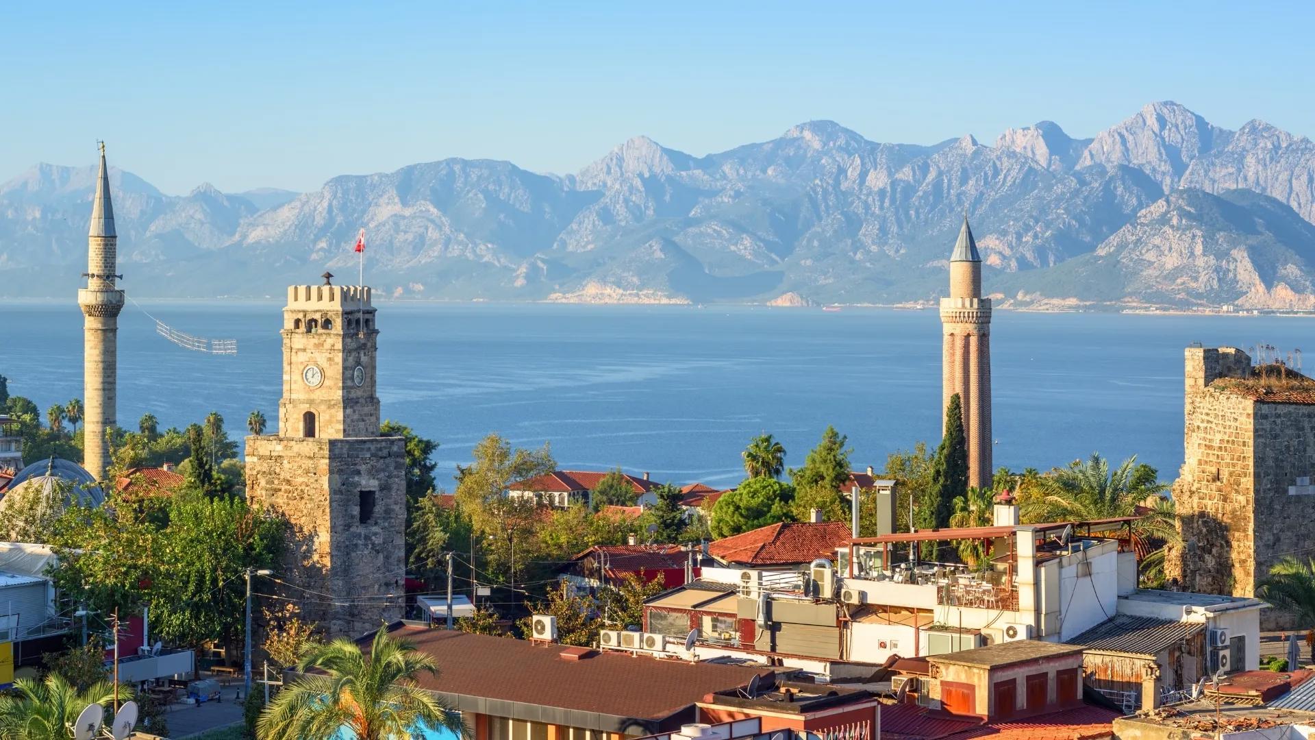 Antalya Old Town skyline with the Clock Tower, historic minarets, red rooftops, and the Taurus Mountains in the background.